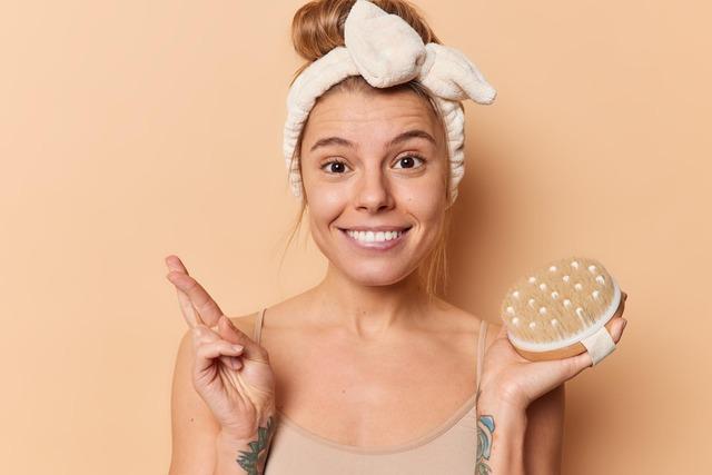 happy-young-woman-holds-body-brush-keeps-fingers-crossed-hopes-good-luck-undergoes-beauty-skin-treatment-procedures-takes-care-herself-wears-headband-casual-t-shirt-poses-indoors_273609-62054
